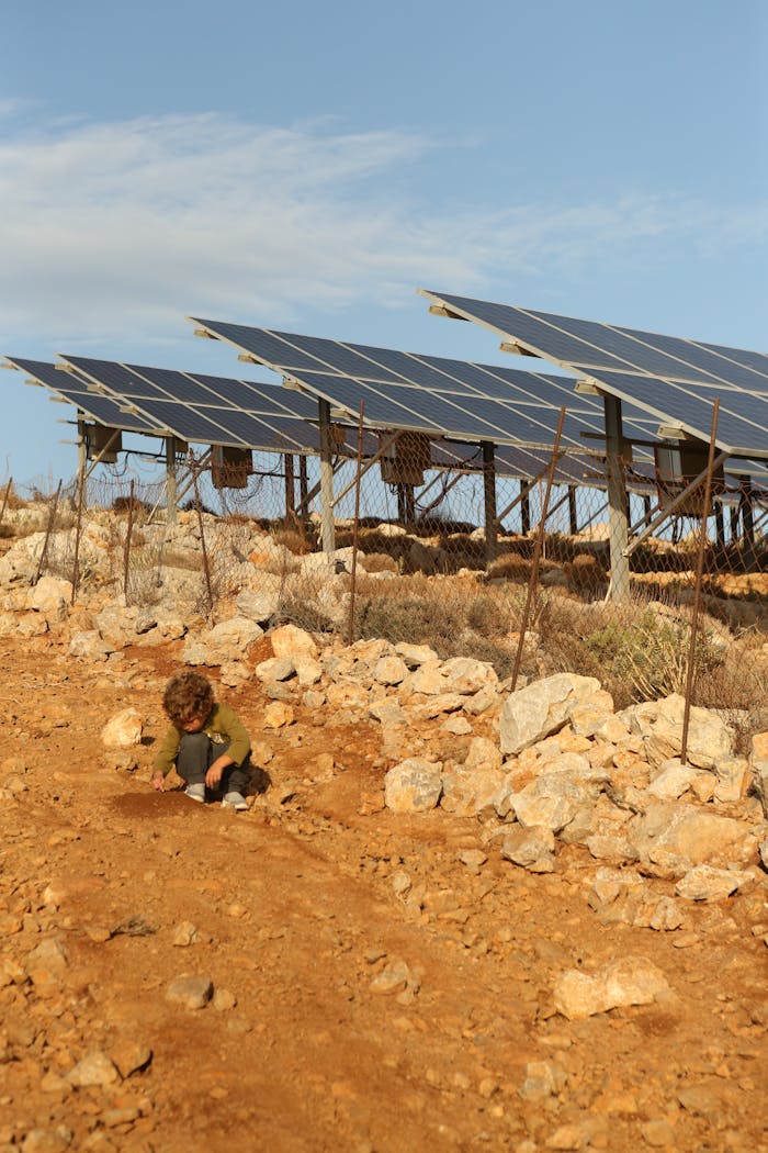 A young child playing on a rocky terrain near solar panels in Greece under a clear sky.